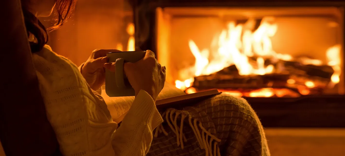 Vi ser litt av en person som sitter med en kopp og en bok foran en peis med ved som brenner