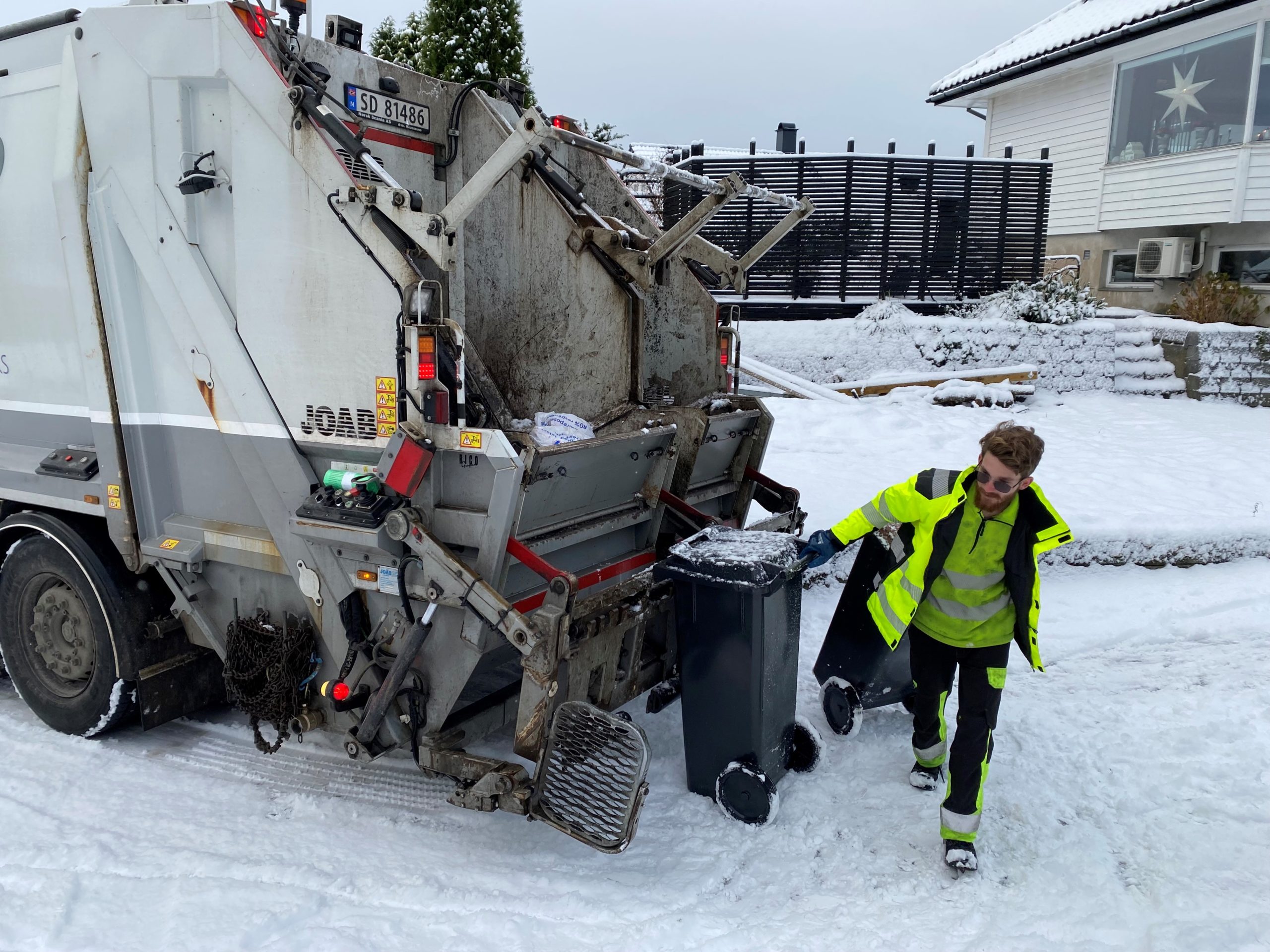 Renovatør som trekker en avfallsbeholder som er tømt i renovasjonsbil. Det er snø.
