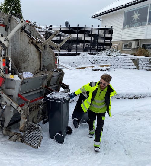 Renovatør som trekker en avfallsbeholder som er tømt i renovasjonsbil. Det er snø.