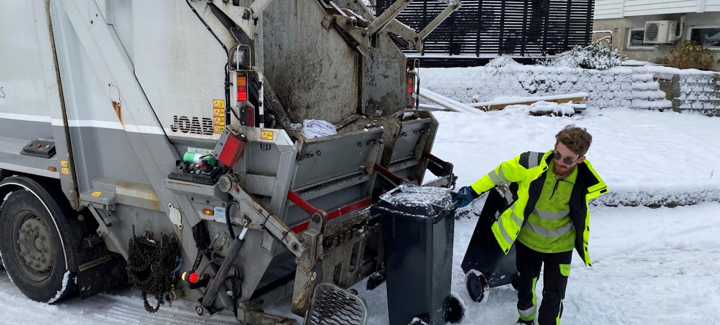 Renovatør som trekker en avfallsbeholder som er tømt i renovasjonsbil. Det er snø.