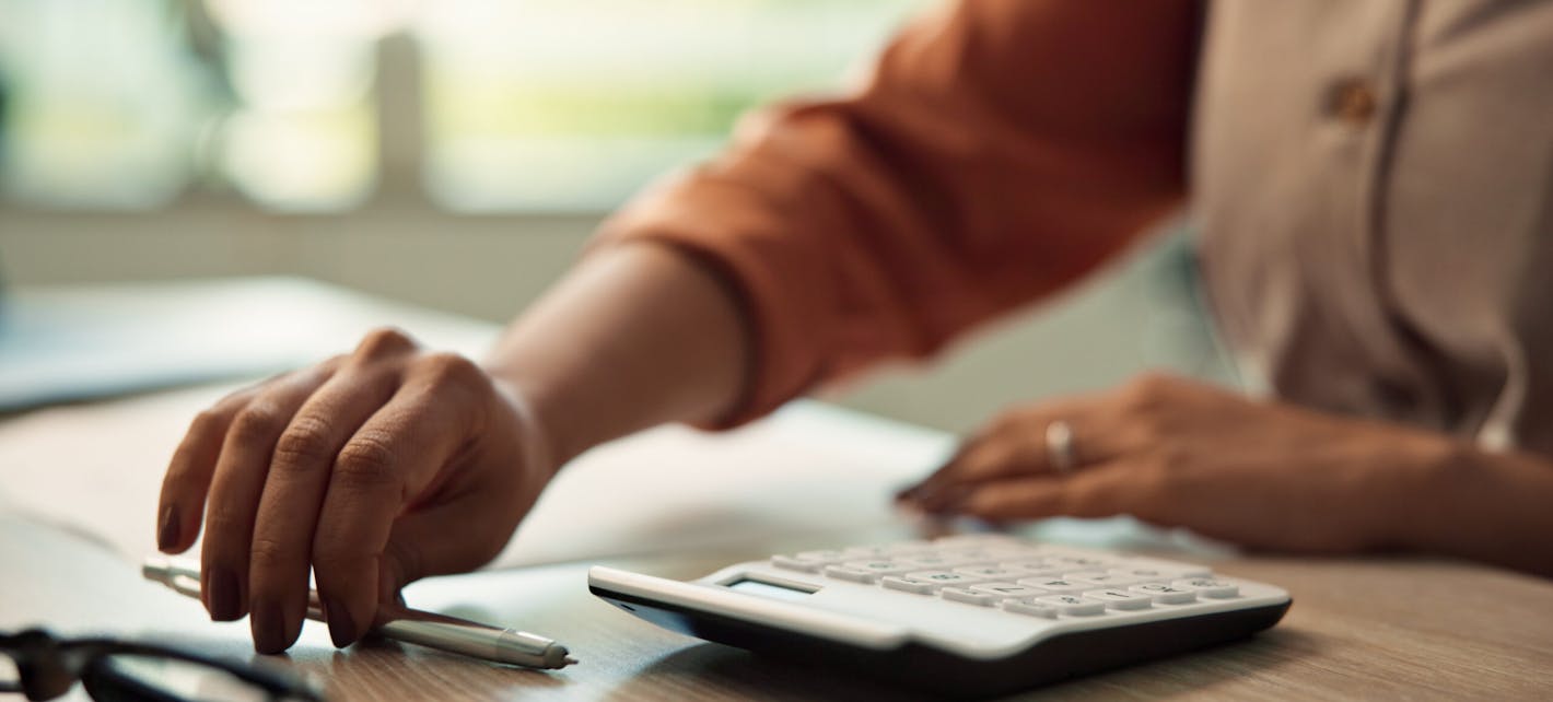 Illustrasjonsbilde som viser en kalkulator og hendene til noen som sitter og jobber med den og litt papirer